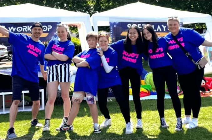 a team of youngsters and 2 adults posing for a picture in a park in Edinburgh while smiling and wearing jojos parties t-shirts.